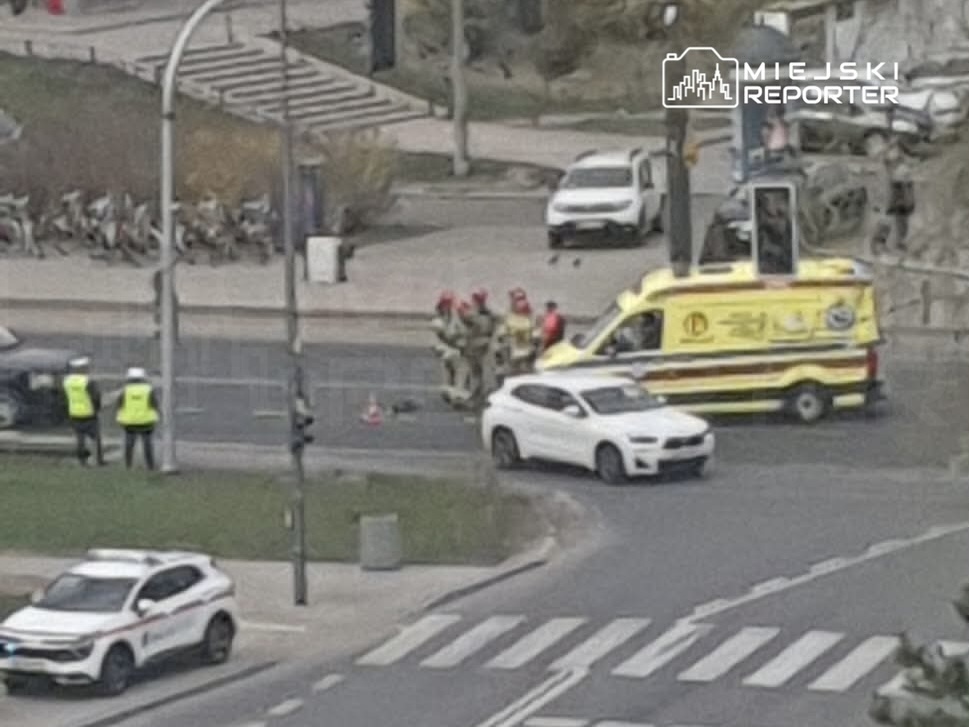 Grupa strażaków w mundurach z czerwonymi hełmami pomaga przy żółtej karetce na skrzyżowaniu ulic.