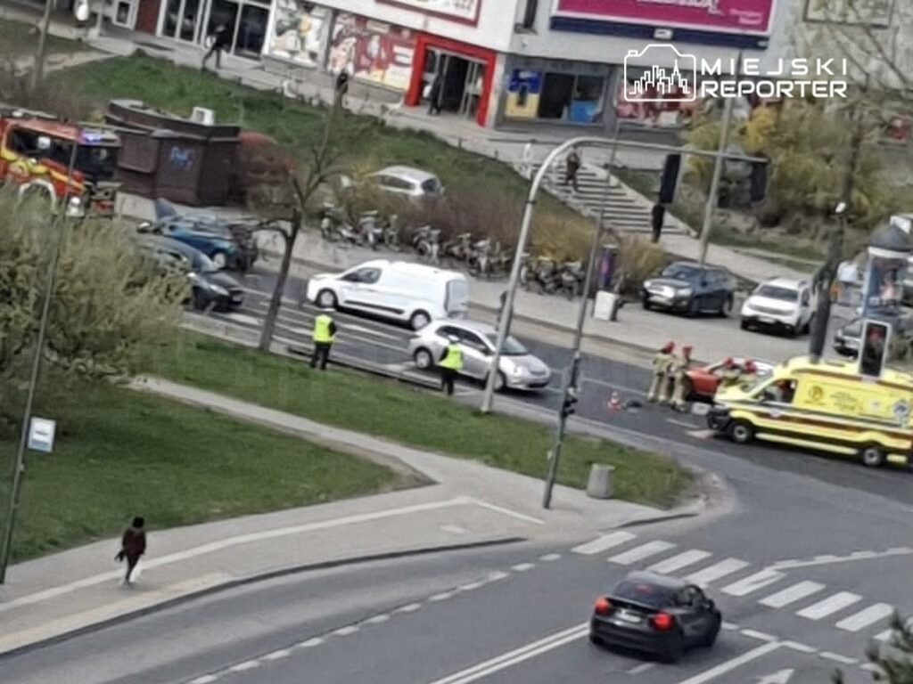 Na skrzyżowaniu stoi biała karetka, obok niej pracują strażacy. W tle widoczne są samochody oraz przechodnie.