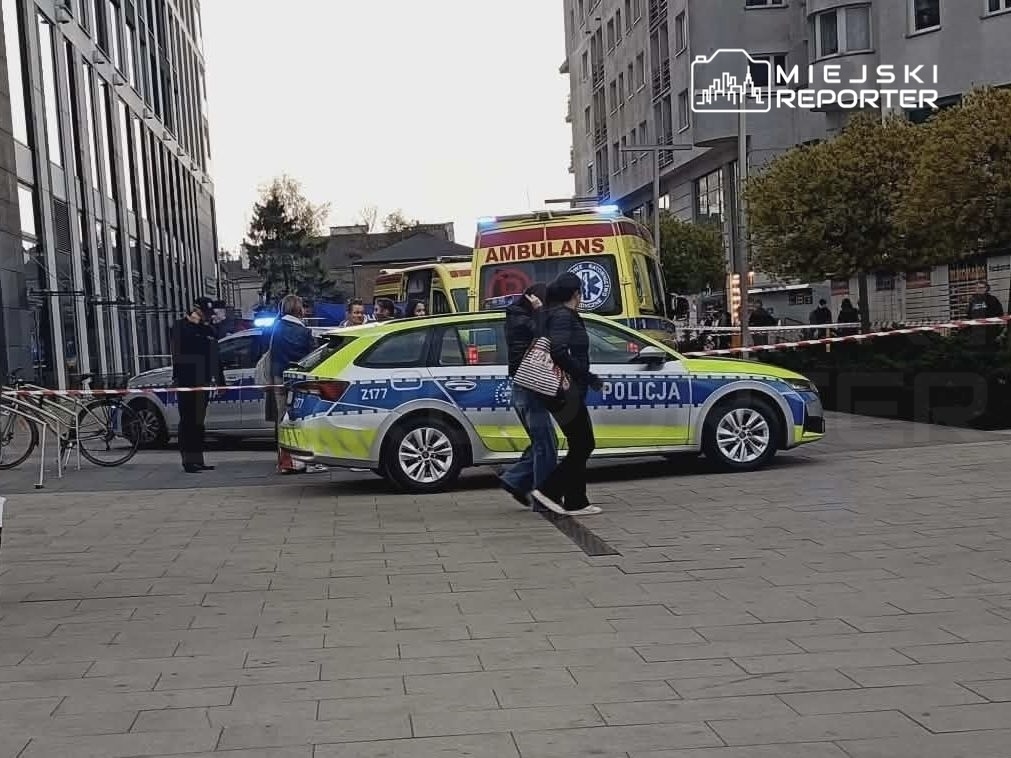 Grupa osób rozmawia w pobliżu radiowozu policji i karetki, otoczona taśmą zabezpieczającą.