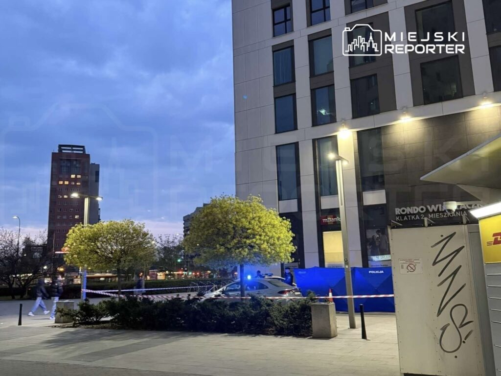 Policja zabezpiecza teren przy nowoczesnym budynku, w tle widać drzewa i oświetlenie uliczne.