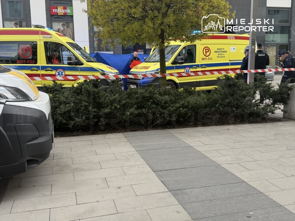 Dwa karetki pogotowia medycznego zaparkowane w pobliżu, obok policjantów zabezpieczających teren.