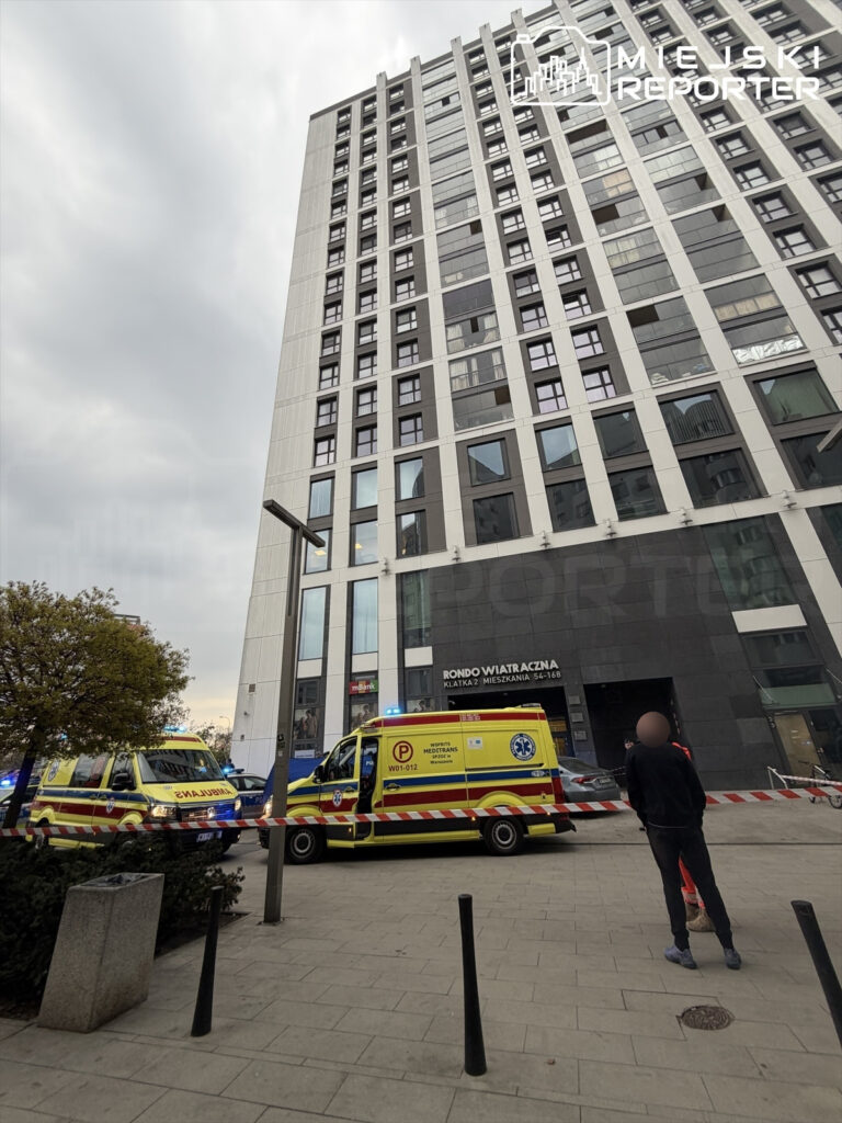 Karetka pogotowia zaparkowana przed nowoczesnym biurowcem, obok mężczyzna obserwujący sytuację.