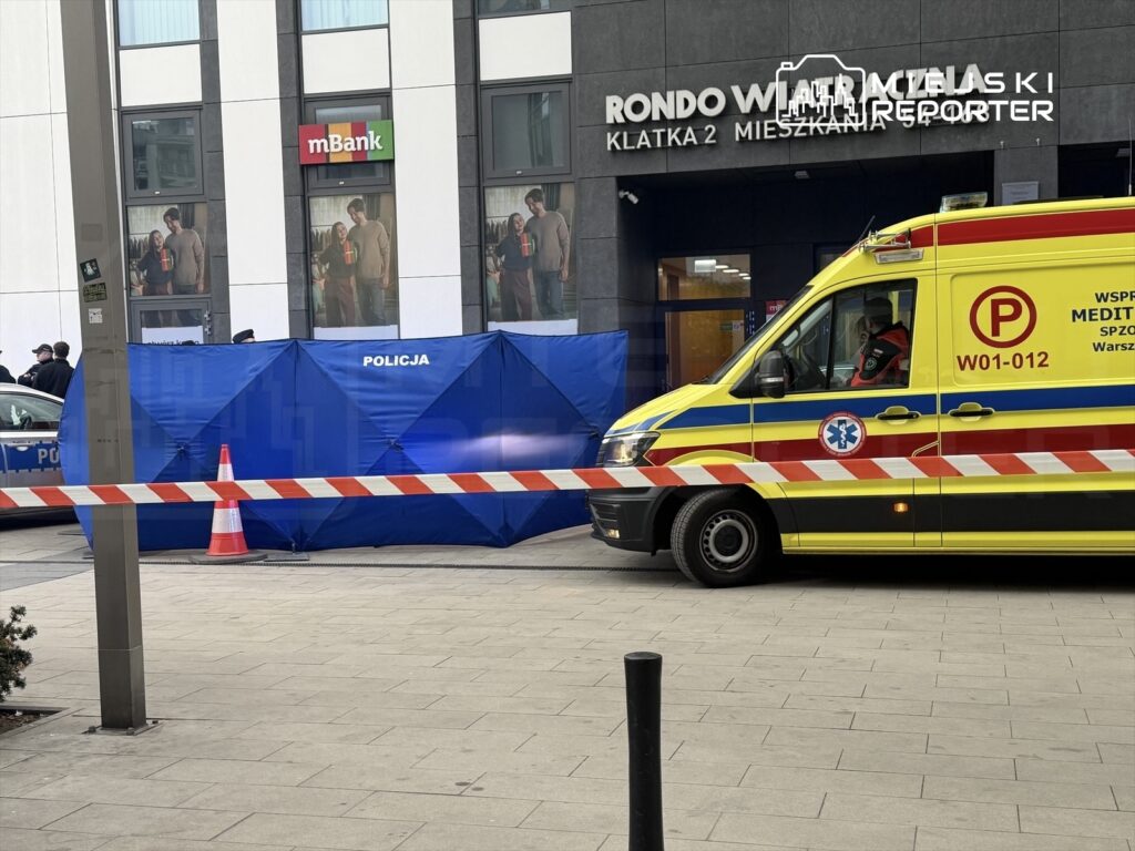 Policjanci zabezpieczają teren przy nowoczesnym biurowcu, obok stoi ambulans z włączonymi sygnałami.