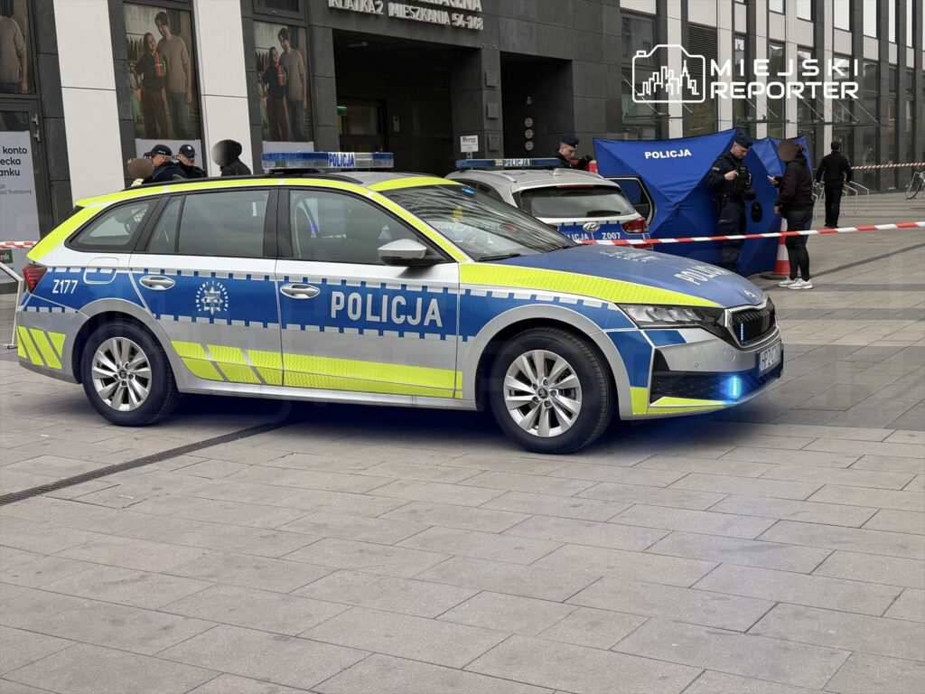 Policyjny samochód osobowy z niebiesko-żółtym oznakowaniem stoi na chodniku, obok grupy funkcjonariuszy rozmawiających przy namiocie.