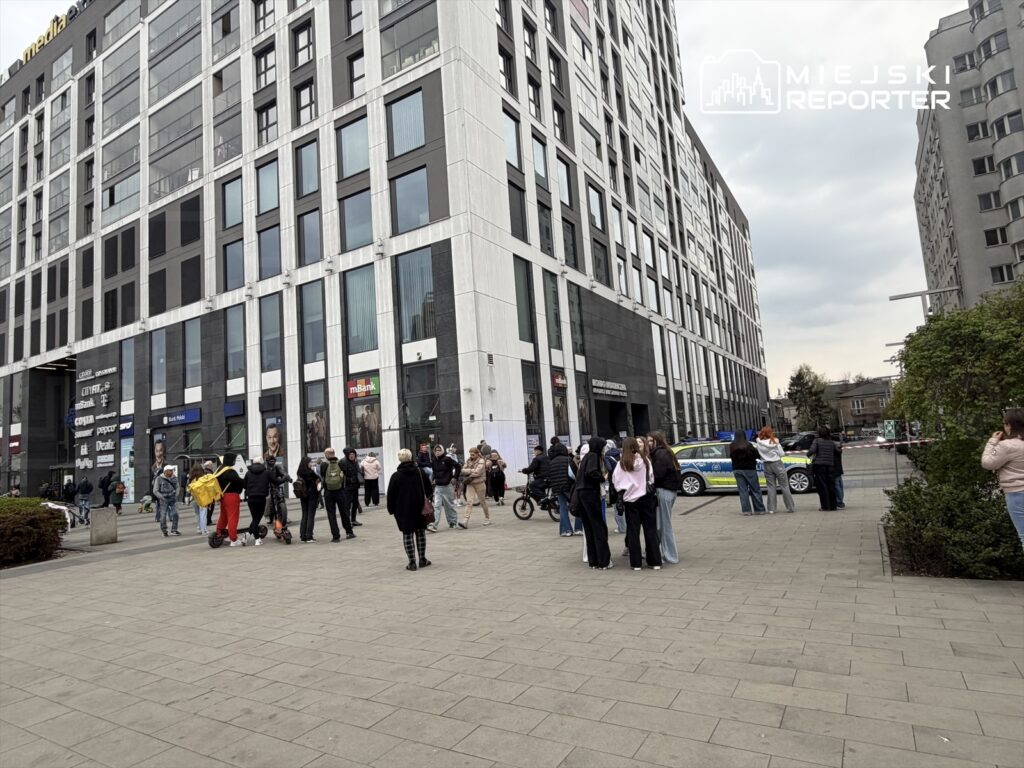 Grupa ludzi gromadzi się przed nowoczesnym biurowcem, niektórzy na hulajnogach, a w tle widoczny radiowóz.