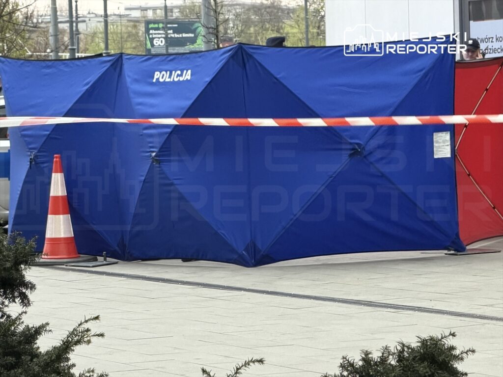 Niebieski namiot policyjny z napisem "POLICJA" ustawiony na placu, otoczony taśmą zabezpieczającą i pachołkami.