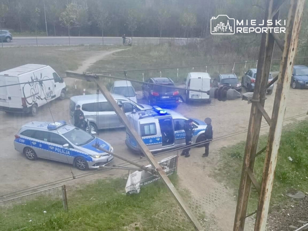 Policjanci rozmawiają obok radiowozu na terenie parkingu z zaparkowanymi pojazdami.