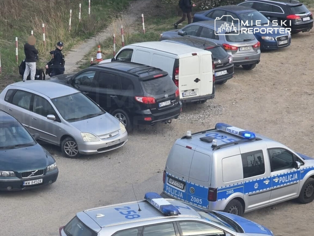 Policjant rozmawia z osobą stojącą obok zaparkowanych samochodów na terenie zarośniętym trawą.