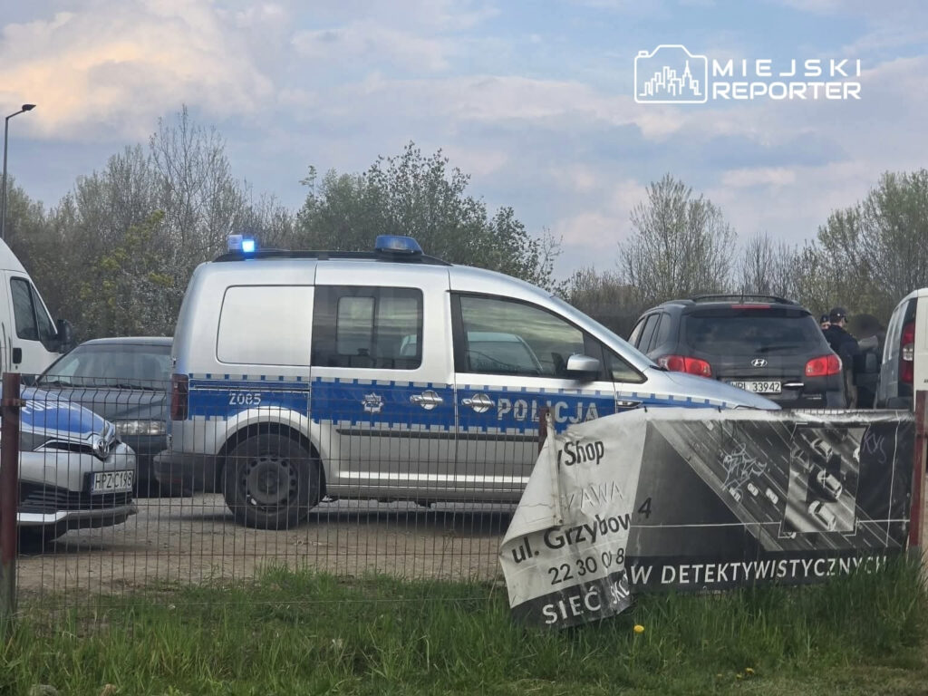 Policyjny radiowóz z włączonymi sygnałami świetlnymi stoi na parkingu, obok zaparkowanych samochodów.
