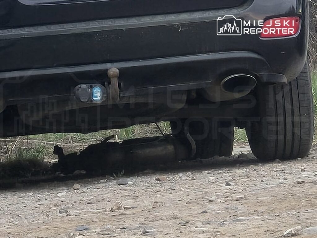 Czarny samochód osobowy zaparkowany na szutrowej drodze, z widocznym uszkodzonym układem wydechowym.