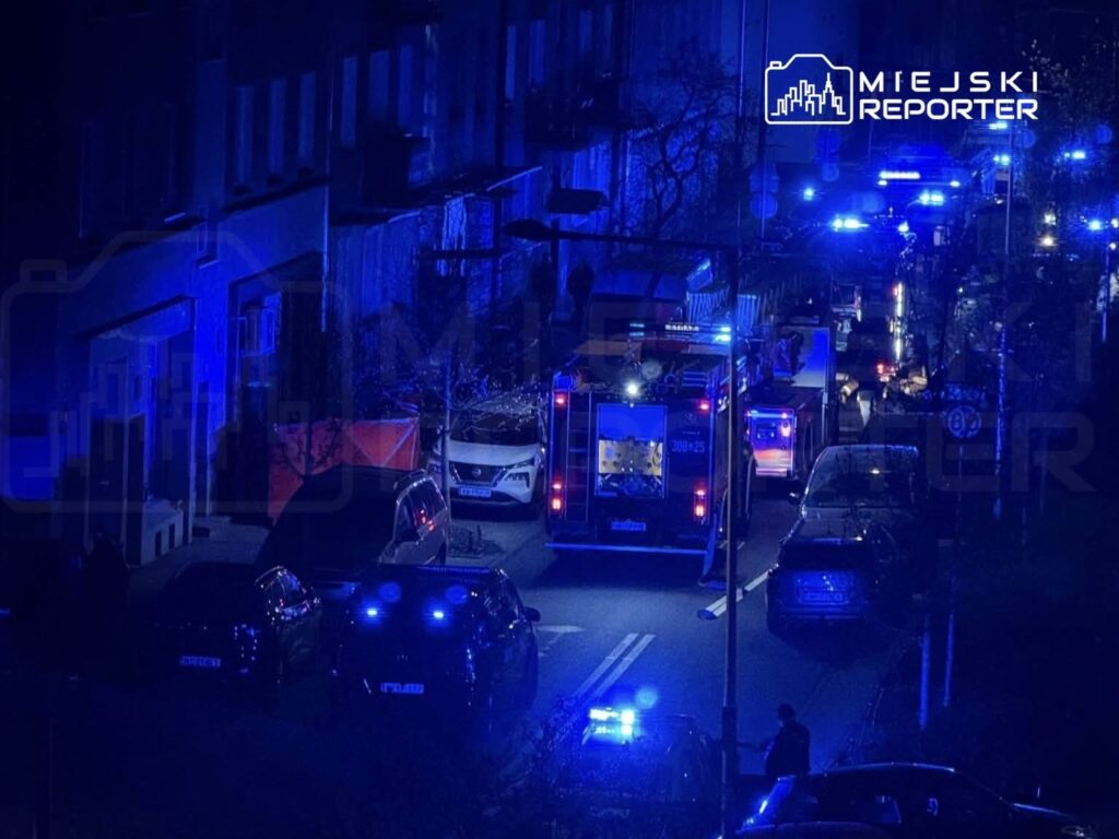 W nocy na ulicy widać pojazdy ratunkowe z włączonymi niebieskimi światłami oraz grupę ludzi w otoczeniu.