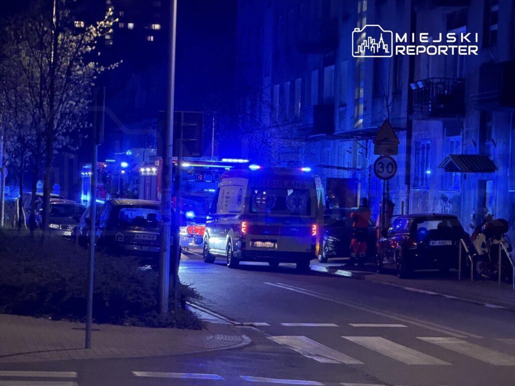 W nocy widać karetki i pojazdy straży pożarnej z włączonymi sygnałami świetlnymi, zaparkowane przy ulicy.