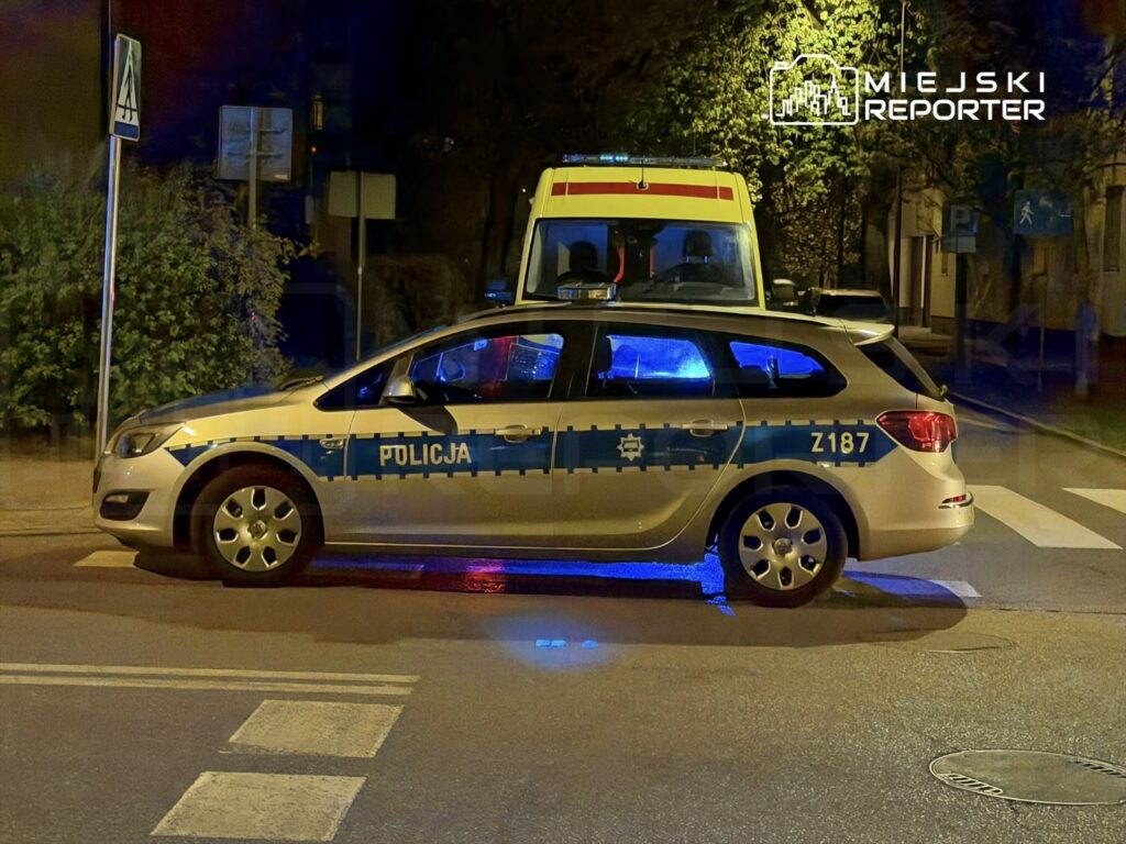 Radiowóz policyjny z niebieskimi światłami zatrzymany na skrzyżowaniu w oświetlonej ulicy nocą.