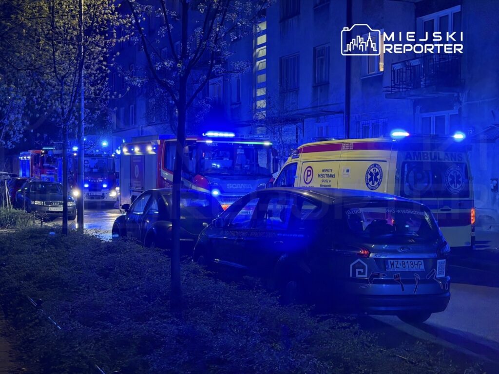 W nocy na ulicy stoją pojazdy ratunkowe, w tym straż pożarna i karetka, oświetlone niebieskimi sygnalizacjami.