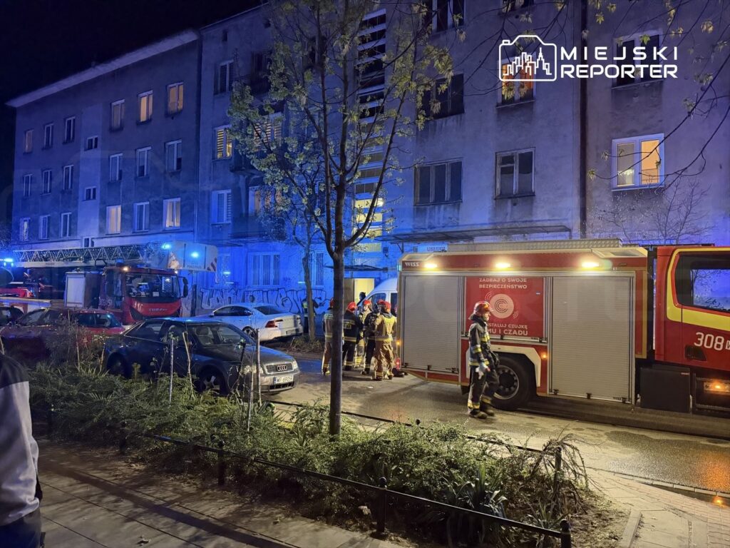 Strażacy w mundurach pracują przy wozie strażackim pod oświetlonym budynkiem mieszkalnym w nocy.