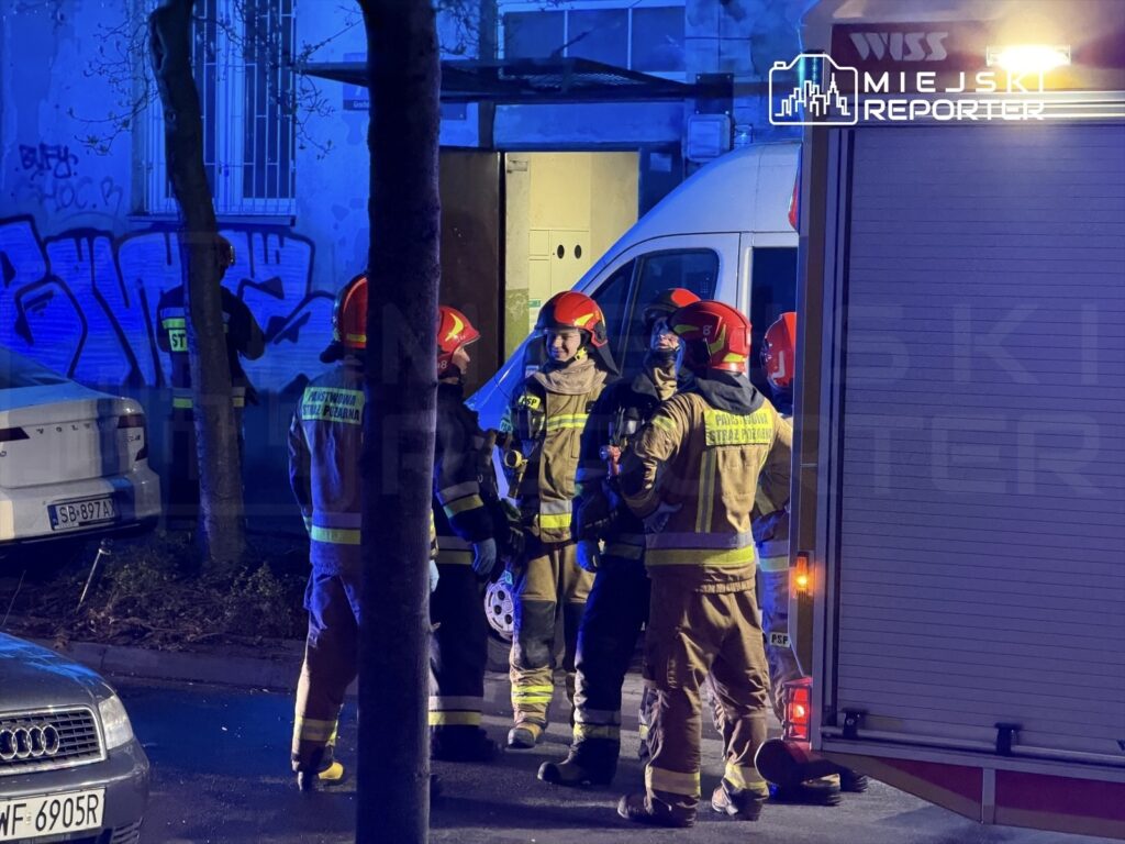Grupa strażaków w hełmach omawia sytuację w nocy przy budynku z graffiti, w tle widoczny wóz strażacki.