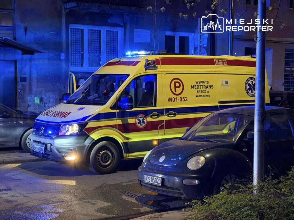 Karetka pogotowia z włączonymi sygnałami świetlnymi stoi na ulicy obok zaparkowanego samochodu.