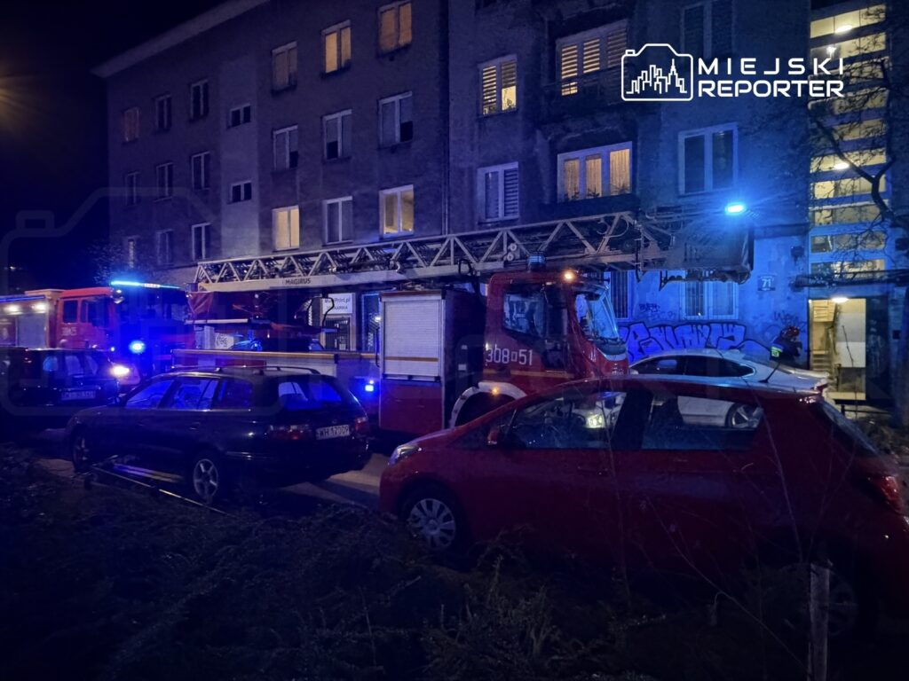 Wozy strażackie z włączonymi sygnałami świetlnymi stoją przed budynkiem mieszkalnym w nocy.