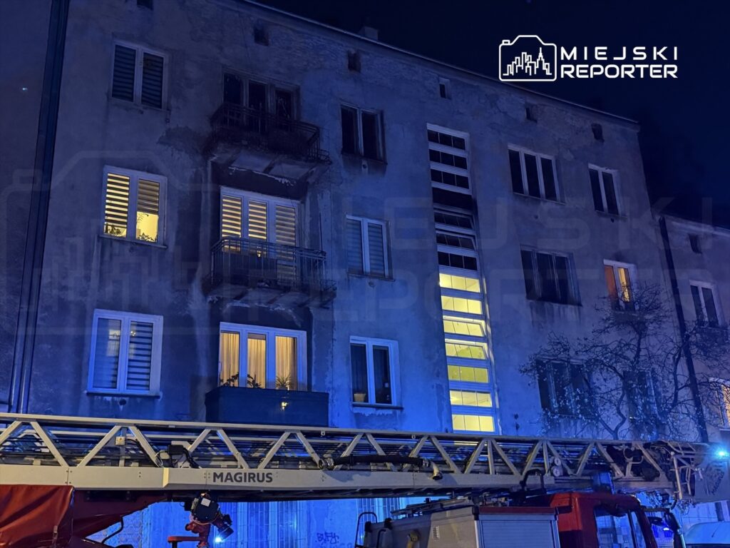 Straż pożarna z drabiną ustawioną przed budynkiem mieszkalnym o szarej elewacji, w nocy z niebieskim oświetleniem.