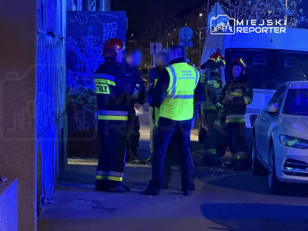 Grupa strażaków w mundurach rozmawia na ulicy, w tle widoczny samochód i oświetlenie w kolorze niebieskim.