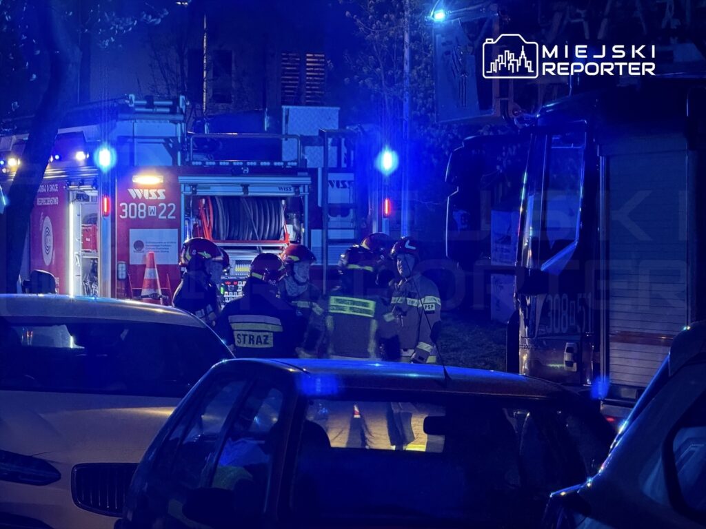 Grupa strażaków w hełmach omawia działania przy wozach strażackich w nocnym miejskim otoczeniu.