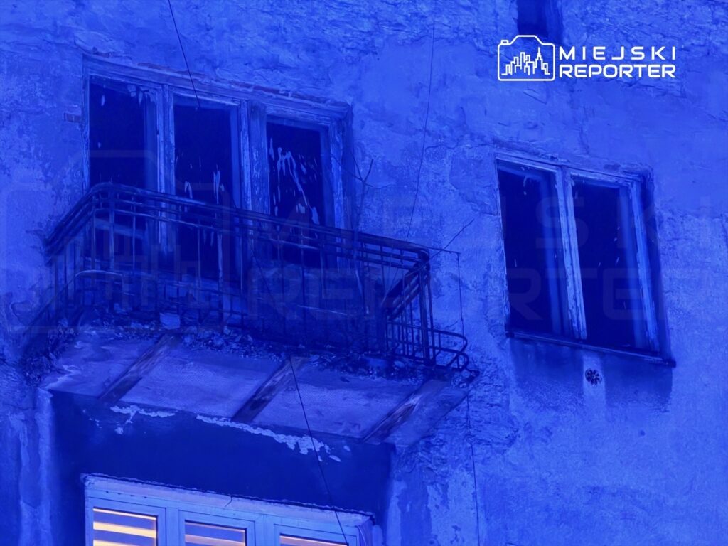 Zniszczony balkon z metalową balustradą i brudnymi oknami w niebieskim świetle.