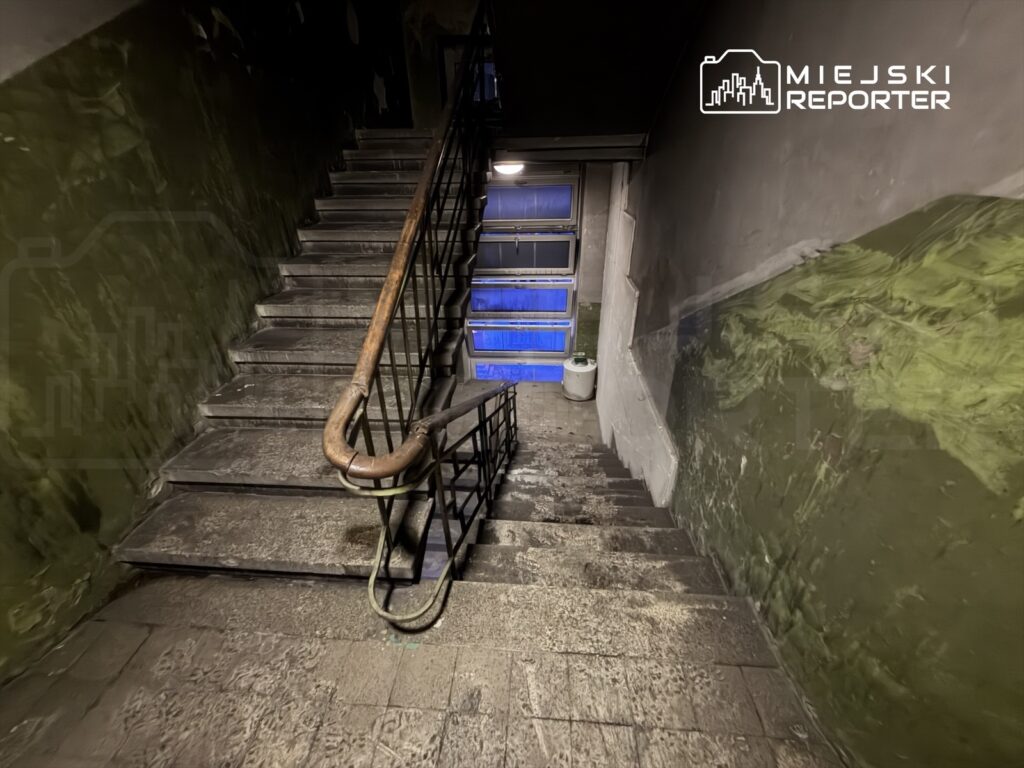 Stare, kamienne schody z metalowym poręczem prowadzące w dół do oświetlonego niebieskim światłem pomieszczenia.