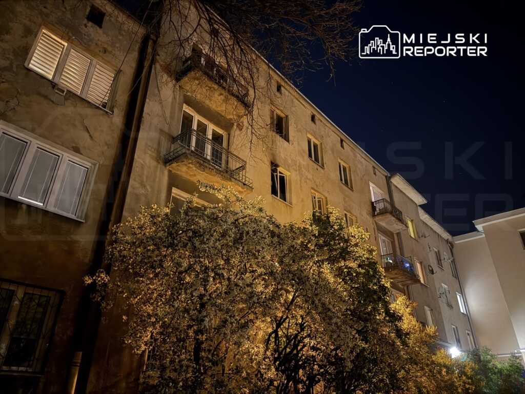 Miejski budynek z balkonami oświetlonymi w nocy, otoczony kwitnącymi drzewami.