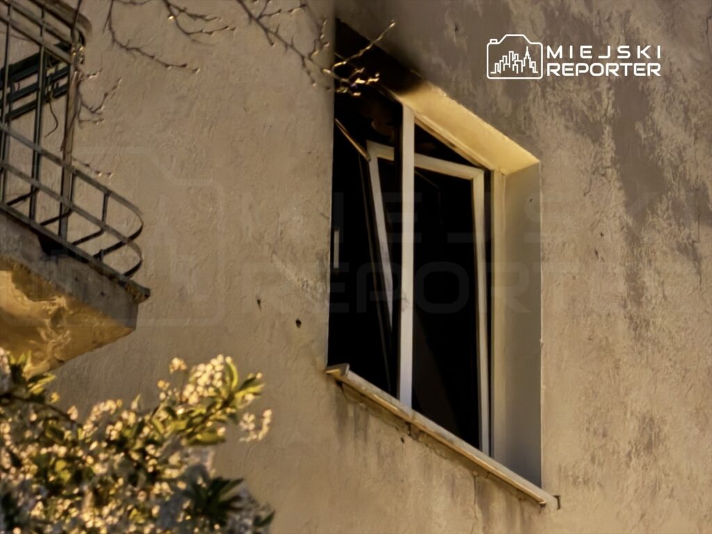 Otwór okienny w ścianie budynku z widocznym metalowym balkonem i roślinnością w tle.