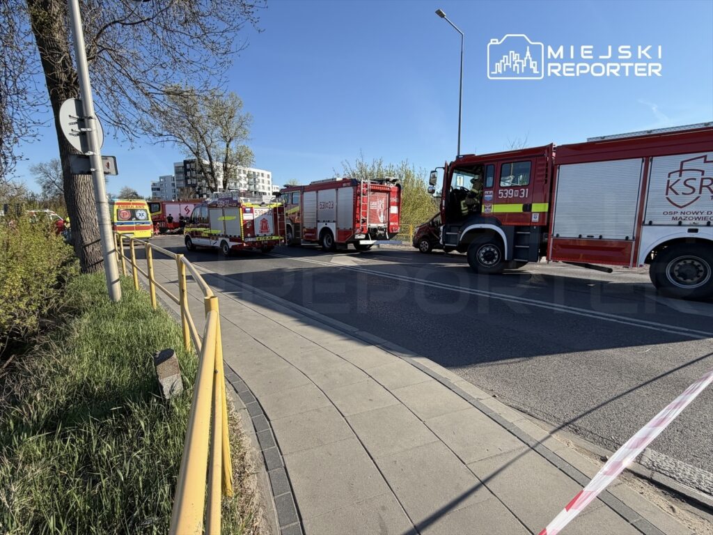 Kilka pojazdów straży pożarnej zaparkowanych na ulicy w pobliżu nowoczesnego budynku mieszkalnego, otoczonego zielenią.