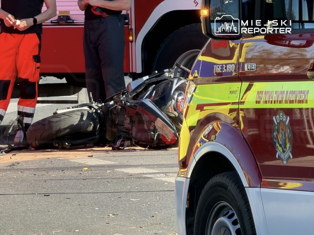 Dwóch ratowników medycznych stoi obok przewróconego motocykla na ulicy, w tle widać wóz strażacki.
