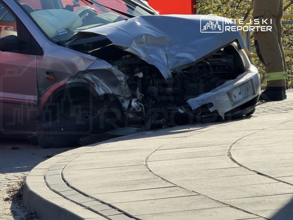 Uszkodzony samochód osobowy z wgniecionym przodem stoi na chodniku, obok widać strażaka w mundurze.