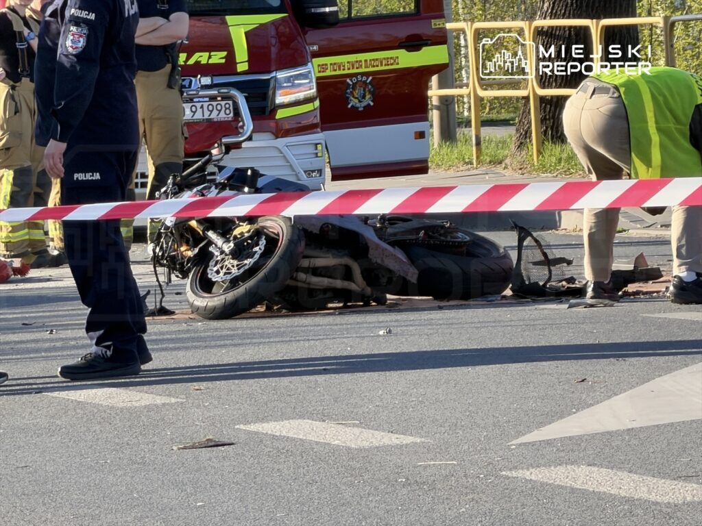 Motocykl leżący na drodze, otoczony przez funkcjonariuszy policji oraz strażaków, z taśmą zabezpieczającą w tle.