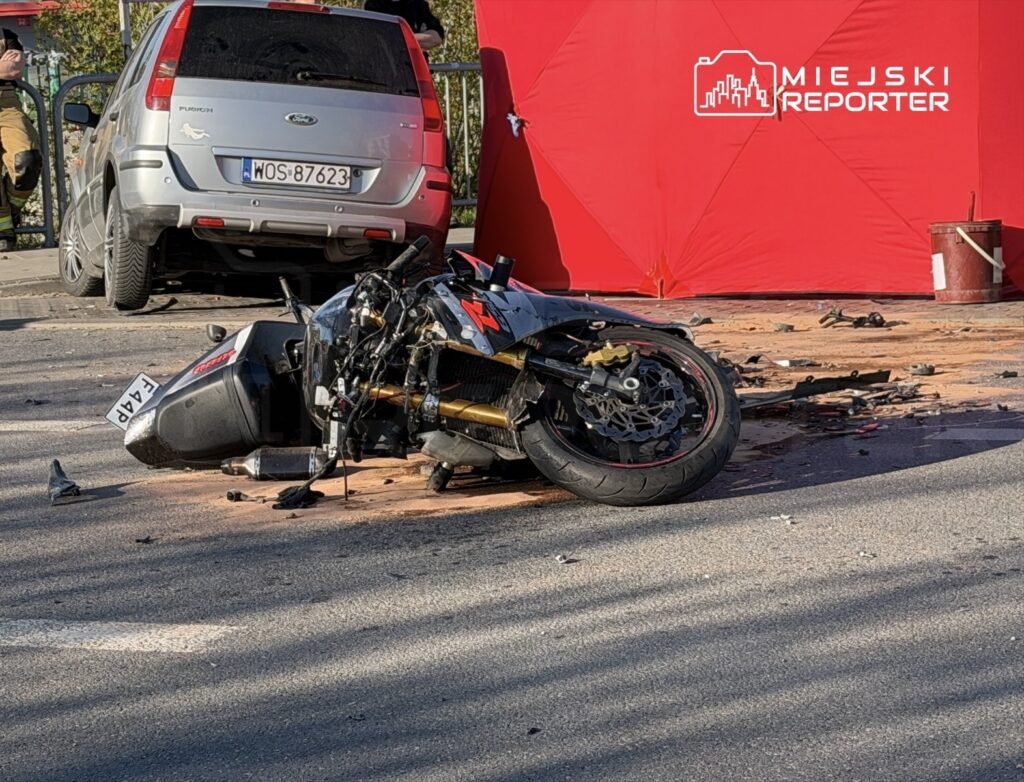 Zniszczony motocykl leży na jezdni obok zaparkowanego samochodu, w tle widoczny czerwony namiot.