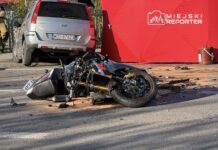 Zniszczony motocykl leży na jezdni obok zaparkowanego samochodu, w tle widoczny czerwony namiot.