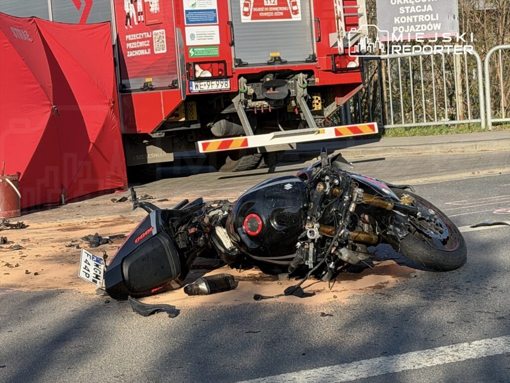 Motocykl leży na drodze, obok rozlane płyny, a w tle widoczna czerwona karetka i namiot zabezpieczający.