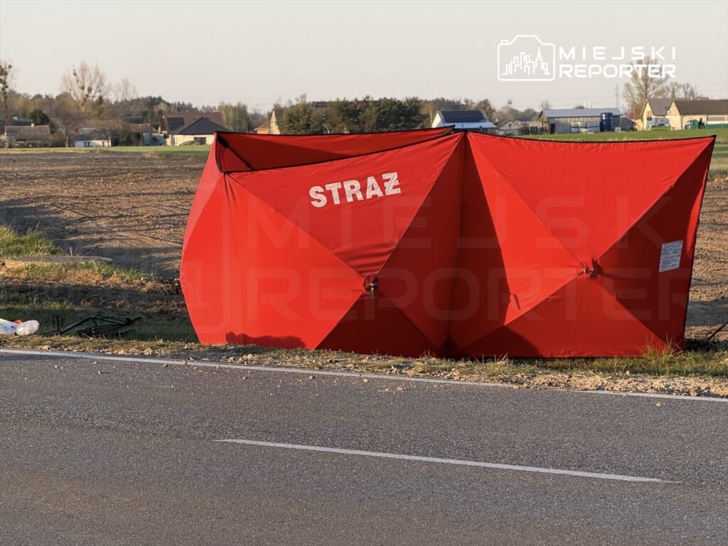 Czerwony namiot z napisem "STRAŻ" ustawiony przy drodze w polu, w tle widoczne domy.