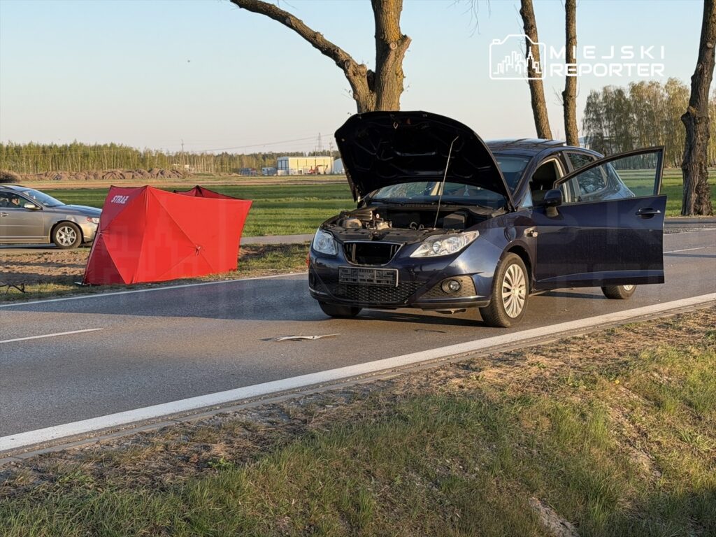 Czarny samochód osobowy z otwartą maską stoi na poboczu drogi, obok czerwony namiot zabezpieczający.