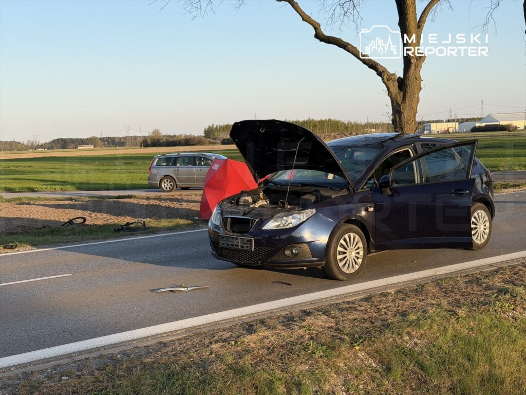 Czarny samochód osobowy z otwartą maską stoi na poboczu drogi, obok leży uszkodzony element. W tle widać drugi pojazd.