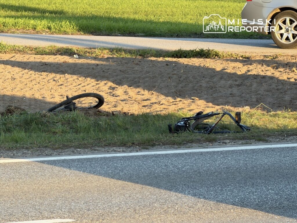 Złamany rower leży na poboczu drogi, obok piaszczystego terenu i trawy.