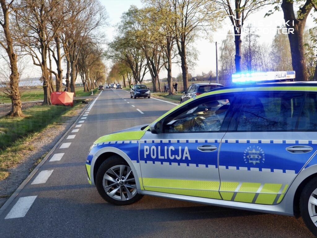 Policyjny radiowóz z niebieskimi światłami zatrzymany przy drodze otoczonej drzewami, obok leży czerwony namiot.