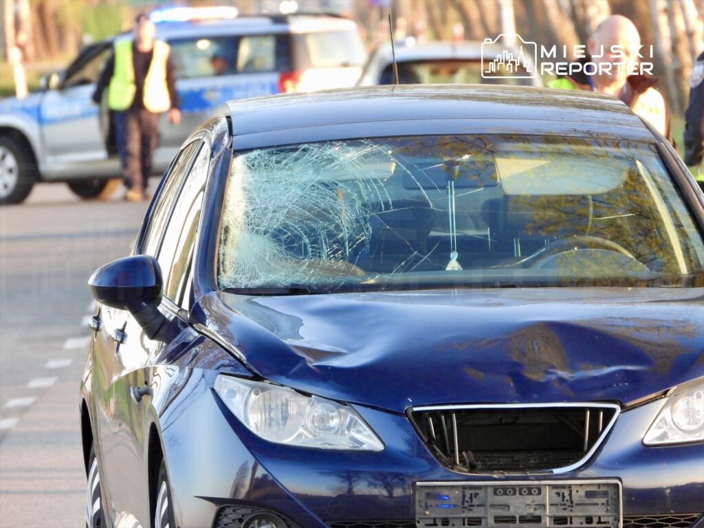 Zniszczony niebieski samochód osobowy z pękniętą szybą przednią, obok policyjny radiowóz i funkcjonariusze.