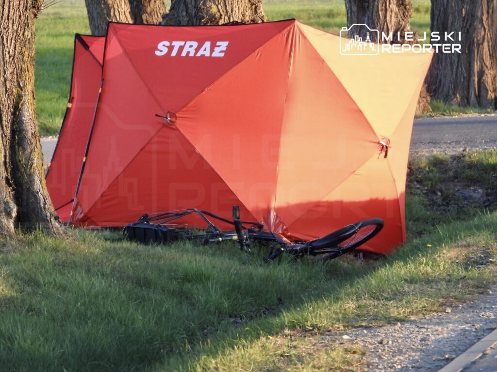 Czerwony namiot z napisem "STRAŻ" ustawiony obok leżącego roweru na trawie przy drodze.