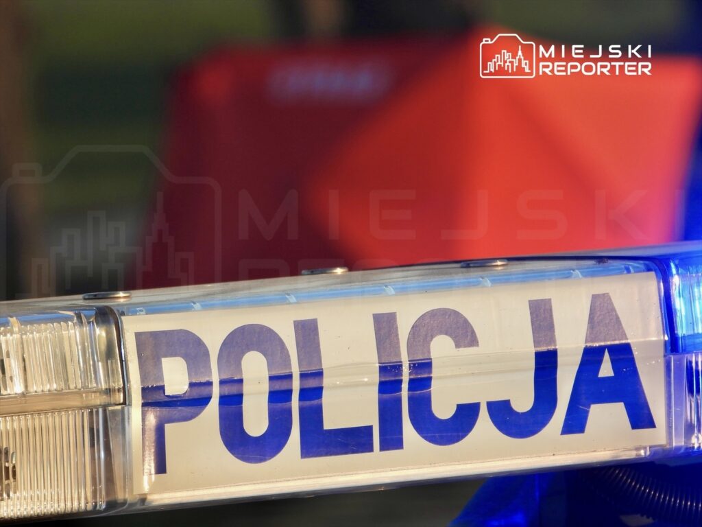 Niebiesko-biała sygnalizacja świetlna z napisem "POLICJA" w tle, w otoczeniu rozmytego czerwonego obiektu.