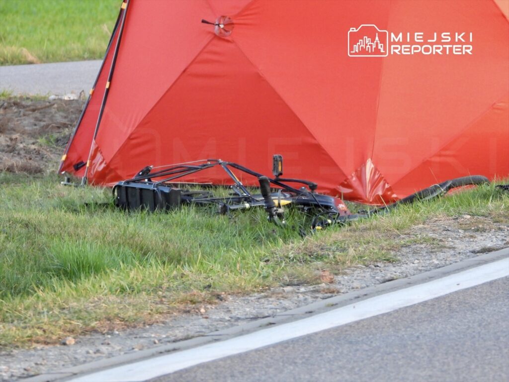 Czerwony namiot rozstawiony na trawie obok drogi, a obok niego leży uszkodzony sprzęt.