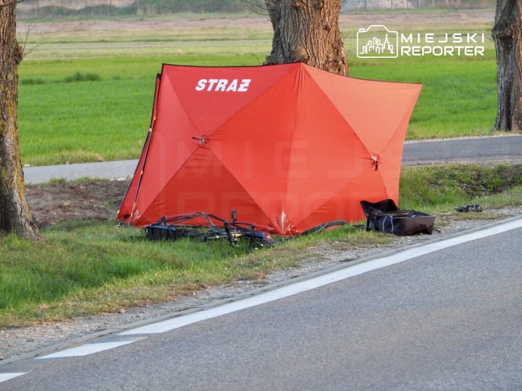 Czerwony namiot z napisem "STRAŻ" ustawiony obok drogi, w otoczeniu zielonych pól i drzew.