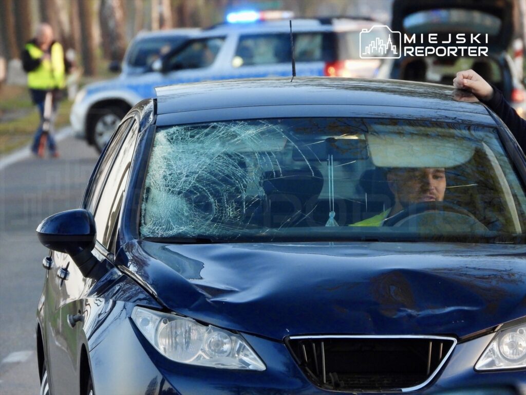 Mężczyzna w samochodzie z rozbitą szybą przednią, obok policyjne pojazdy na drodze.