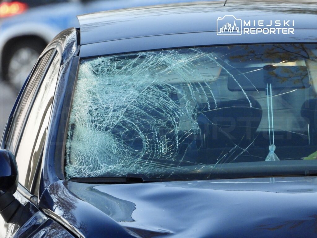 Pęknięta szyba czołowa czarnego samochodu stojącego na parkingu, w tle widoczne inne pojazdy.