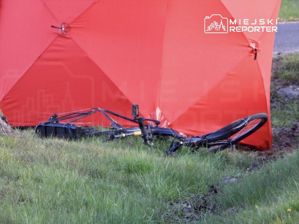 Czarny rower leży na trawie obok czerwonego namiotu w terenie zielonym przy drodze.