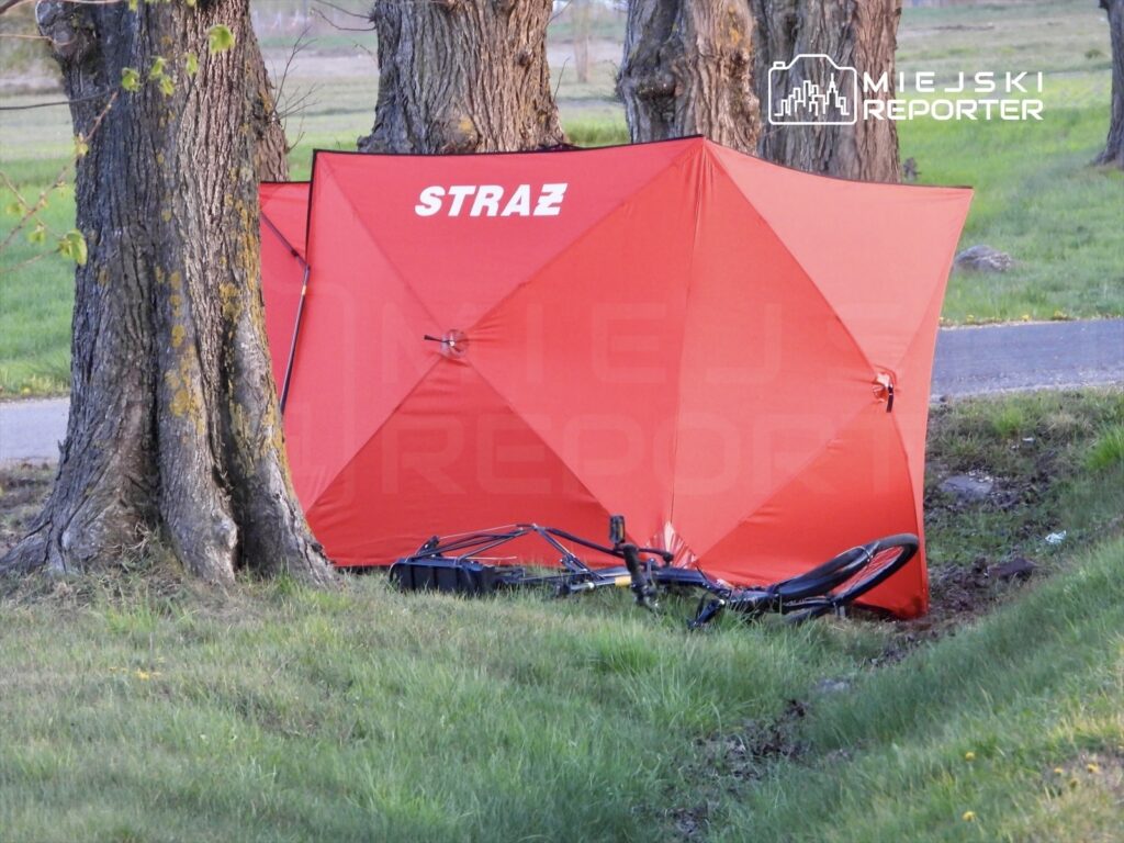 Czerwony namiot strażacki rozstawiony obok leżącego roweru w trawie przy drodze.